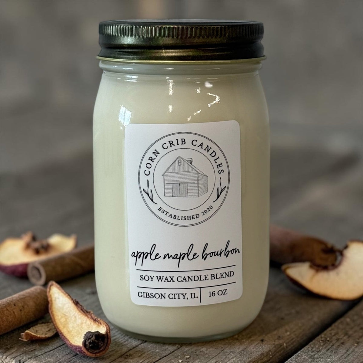 Apple Maple Bourbon 16 oz. candle in a glass jar with a black lid, surrounded by apple slices and cinnamon sticks